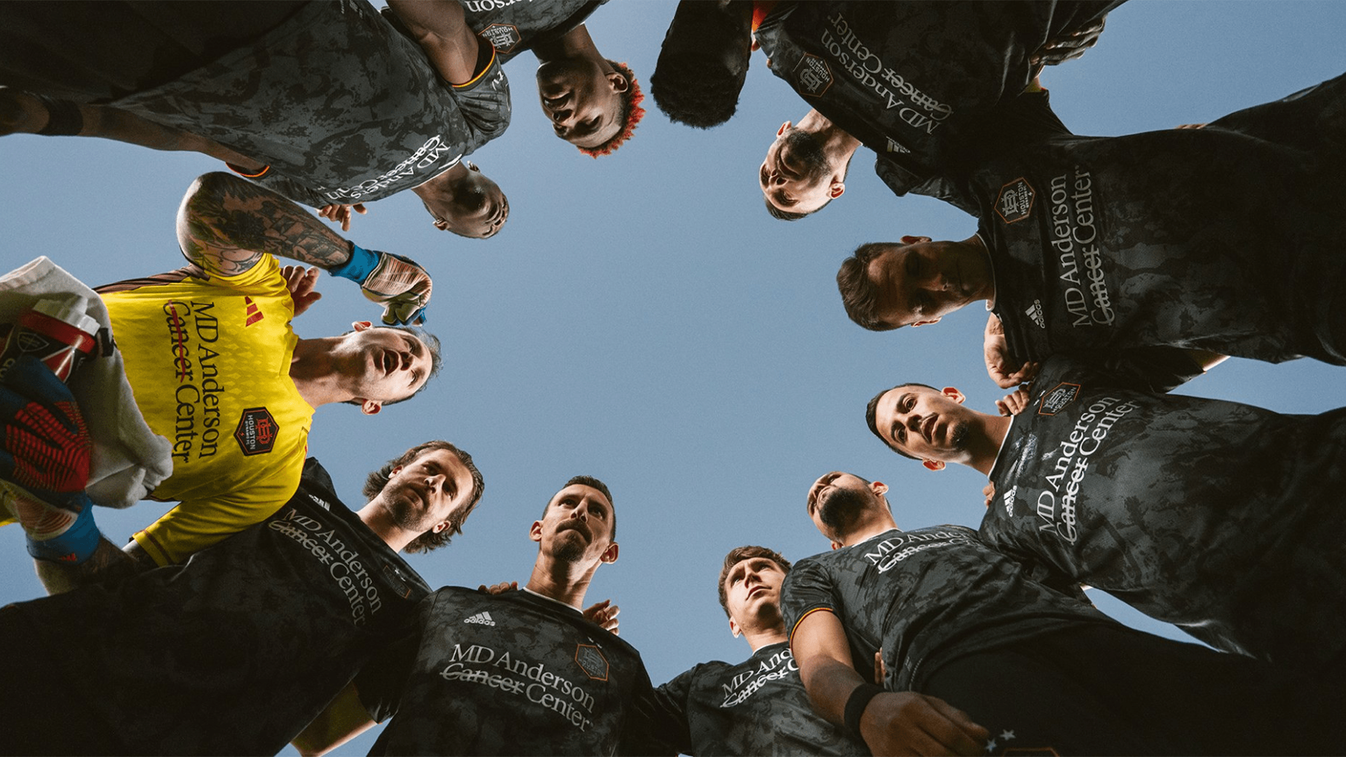 Eleven Houston Dynamo soccer team members huddling together above a low-angle straight-up shot, showcasing unity and determination before a match.