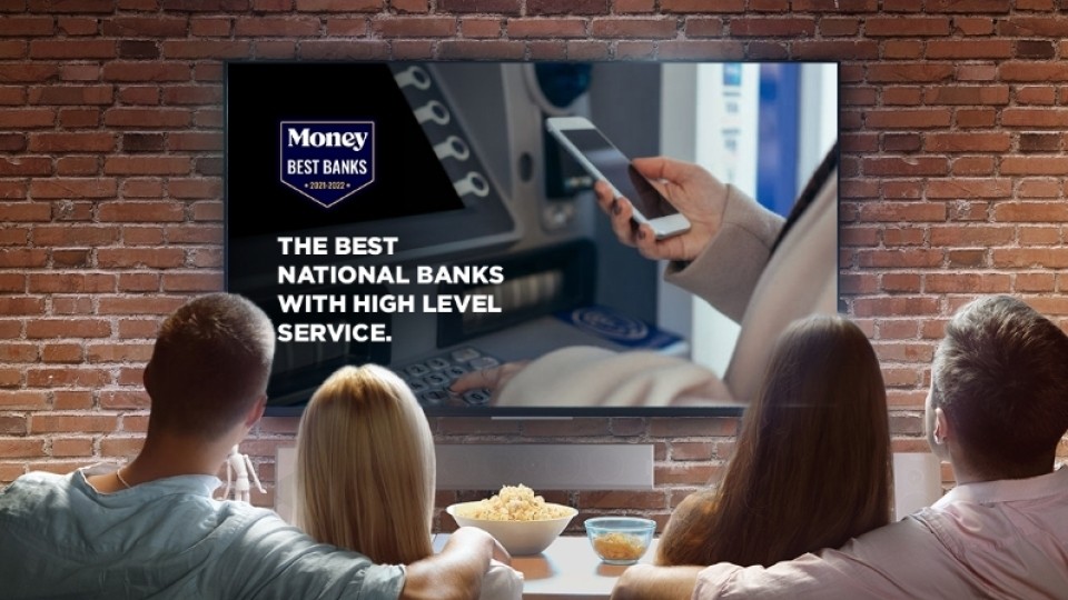 Two couples relax in a living room, sharing a bowl of popcorn while watching a big-screen TV. Visible on screen is a commercial for a local bank employing the Money Best Banks award logo.